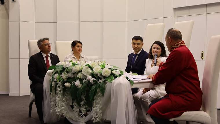 Çiçeklerle süslenmiş masanın arkasında oturan çiftin nikâhını, mikrofonlu bir kadın ve kırmızı cüppeli kişi yürütüyor.