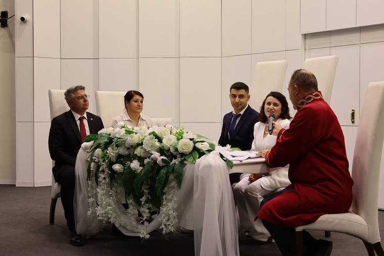 Çiçeklerle süslenmiş masanın arkasında oturan çiftin nikâhını, mikrofonlu bir kadın ve kırmızı cüppeli kişi yürütüyor.