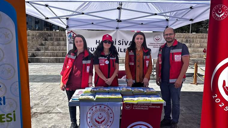 Açık alandaki çadır altında dört kişi, üzerinde broşür ve kitapçıklar bulunan masanın arkasında duruyor.