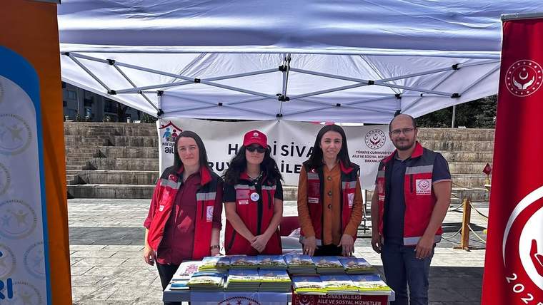 Açık alandaki çadır altında dört kişi, üzerinde broşür ve kitapçıklar bulunan masanın arkasında duruyor.