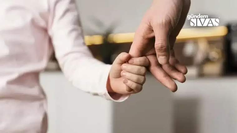 Bir yetişkinin parmağını tutan çocuğun eli, yakın planda açık renkli bir giysi önünde görülüyor.
