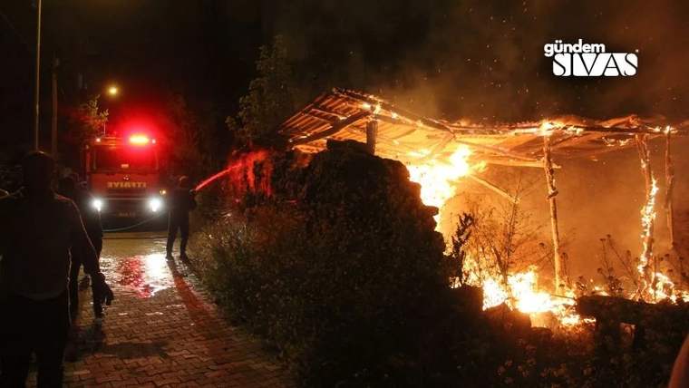 Gece, alevler içindeki tek katlı yapının yanında kırmızı ışıkları yanan itfaiye aracı ve insanlar görülüyor.
