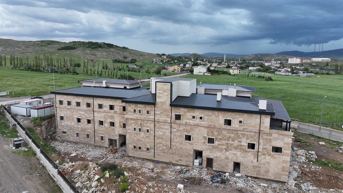 Kırsal alanda, çevresi molozlarla kaplı ve dış cephe çalışmaları süren büyük bir bina görülüyor.