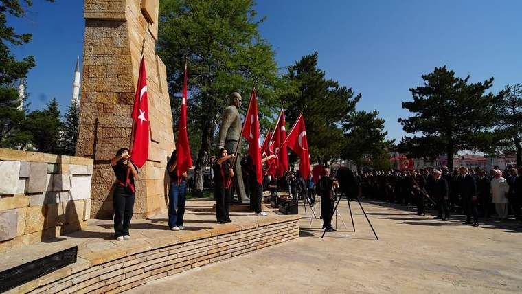 Taş anıt önünde Türk bayrakları taşıyan kişiler ve tören alanında sıralanan kalabalık görülüyor.