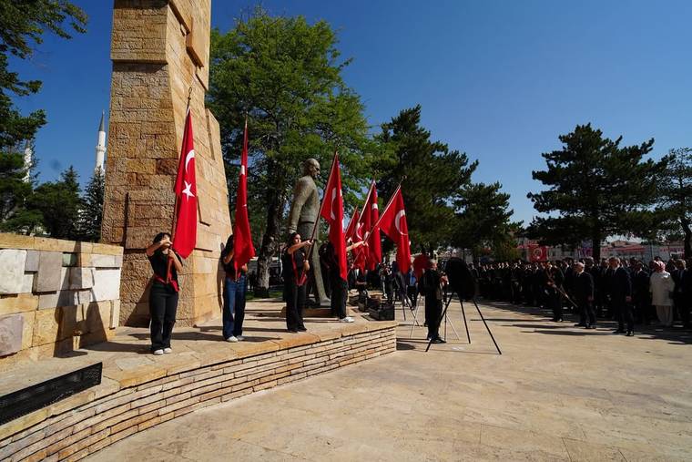 Taş anıt önünde Türk bayrakları taşıyan kişiler ve tören alanında sıralanan kalabalık görülüyor.