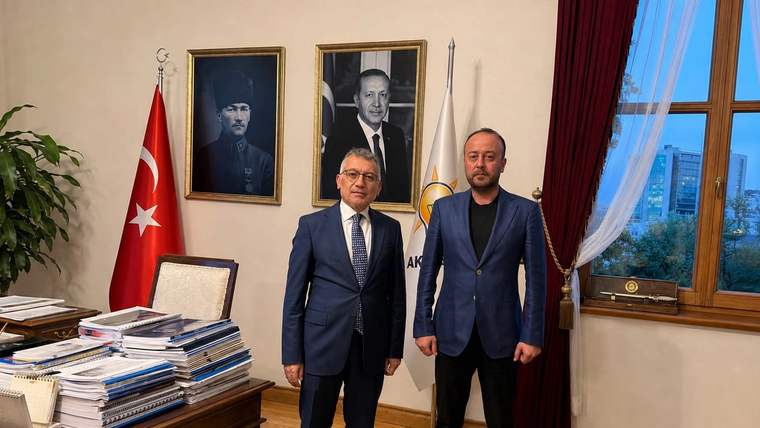 Türk bayrağı ve iki portre önünde takım elbiseli iki kişi, ahşap mobilyalı bir odada duruyor.