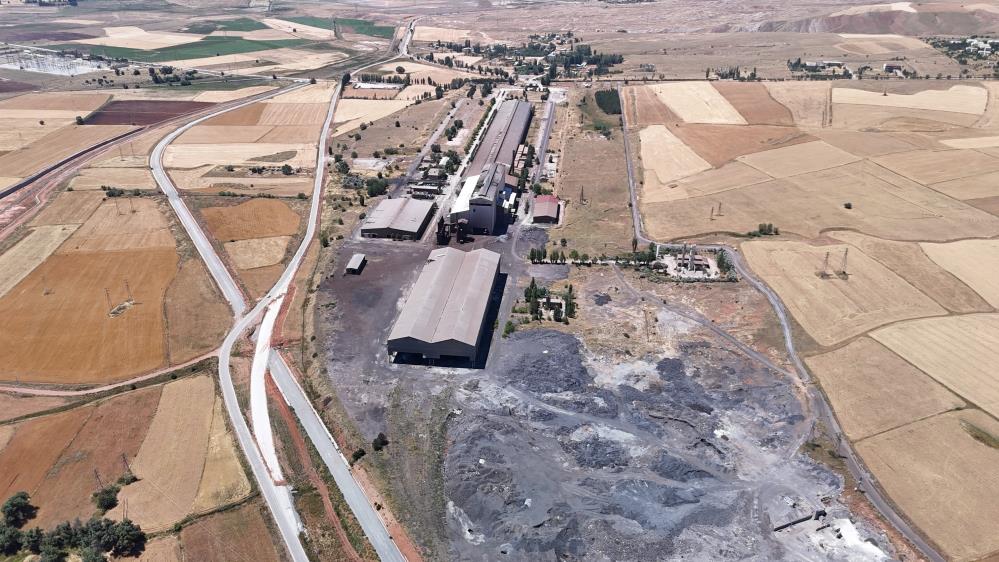 Geniş tarım arazileri arasında, büyük endüstriyel yapılar ve çevresinde gri yığınların bulunduğu bir tesis görülüyor.