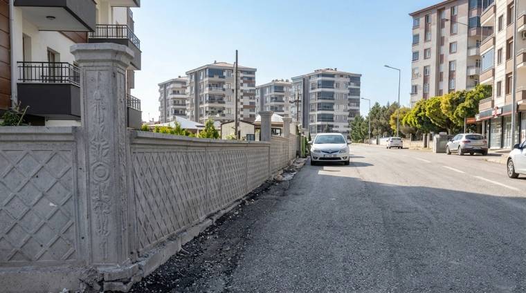 Apartmanların bulunduğu geniş caddede, duvar boyunca asfaltla yol arasında taşlı ve düzensiz bir şerit görülüyor.