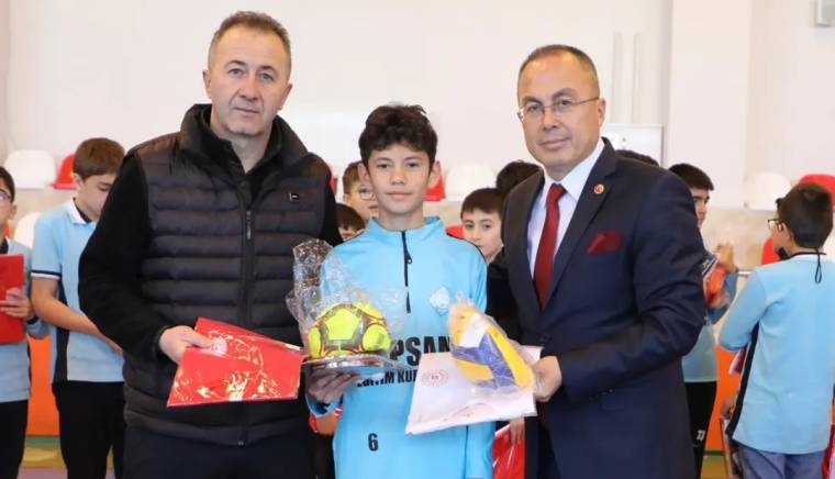 İki kişi, elinde futbol topu tutan bir çocuğa ödül veriyor.