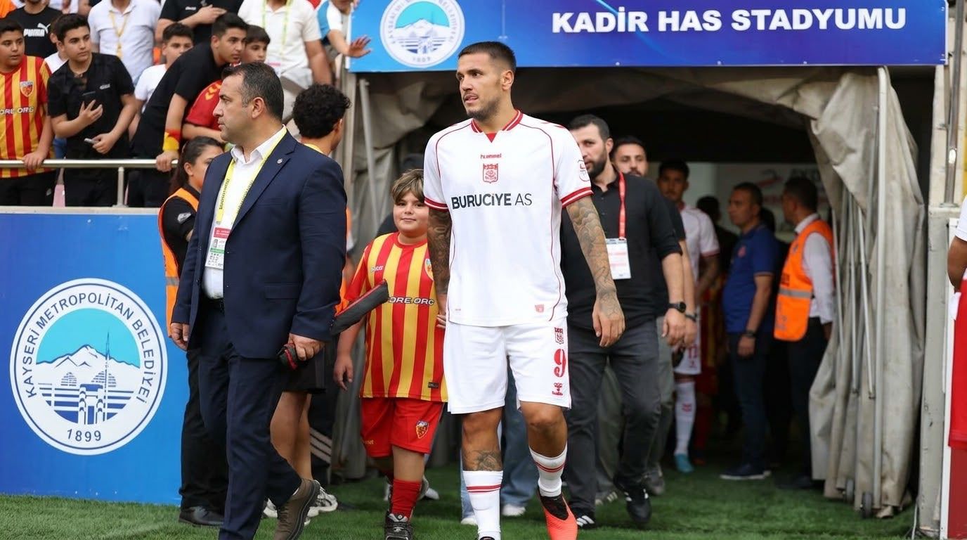 Beyaz formalı bir futbolcu, çocuk ve görevlilerle stadyum tünelinden çıkıyor; üstte Kadir Has Stadyumu tabelası.