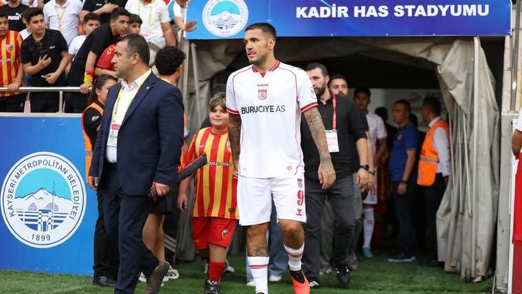 Beyaz formalı bir futbolcu, çocuk ve görevlilerle stadyum tünelinden çıkıyor; üstte Kadir Has Stadyumu tabelası.