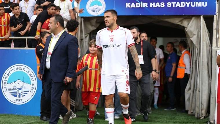 Beyaz formalı bir futbolcu, çocuk ve görevlilerle stadyum tünelinden çıkıyor; üstte Kadir Has Stadyumu tabelası.