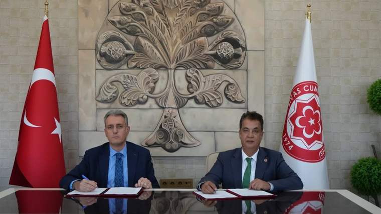 Türk bayrağı ve kurum flamaları önünde iki kişi, masadaki belgeleri imzalarken görülüyor.