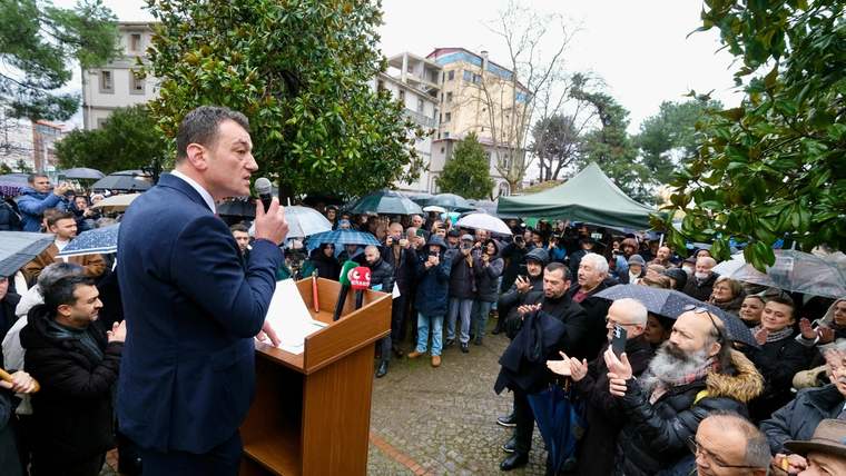 Mikrofonla konuşan bir kişi, yağmurluk ve şemsiyeler taşıyan kalabalığa ahşap kürsüden sesleniyor.