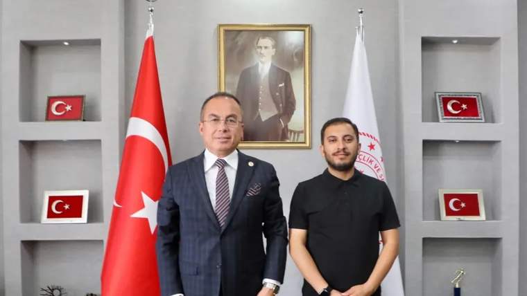 Bir odada iki kişi, Türk bayrağı ve kurum bayrağının önünde, duvardaki portre altında yan yana duruyor.
