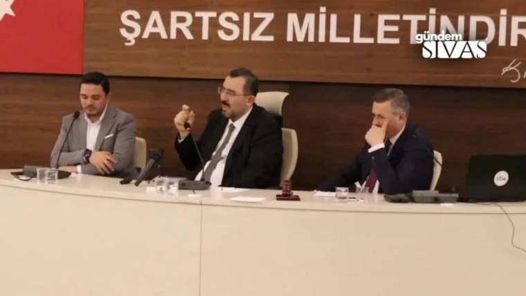 Masada oturan üç kişiden ortadaki kişi mikrofon tutarken, arkadaki panoda “ŞARTSIZ MİLLET...” yazısı görülüyor.