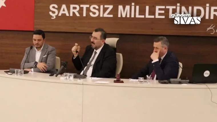 Masada oturan üç kişiden ortadaki kişi mikrofon tutarken, arkadaki panoda “ŞARTSIZ MİLLET...” yazısı görülüyor.
