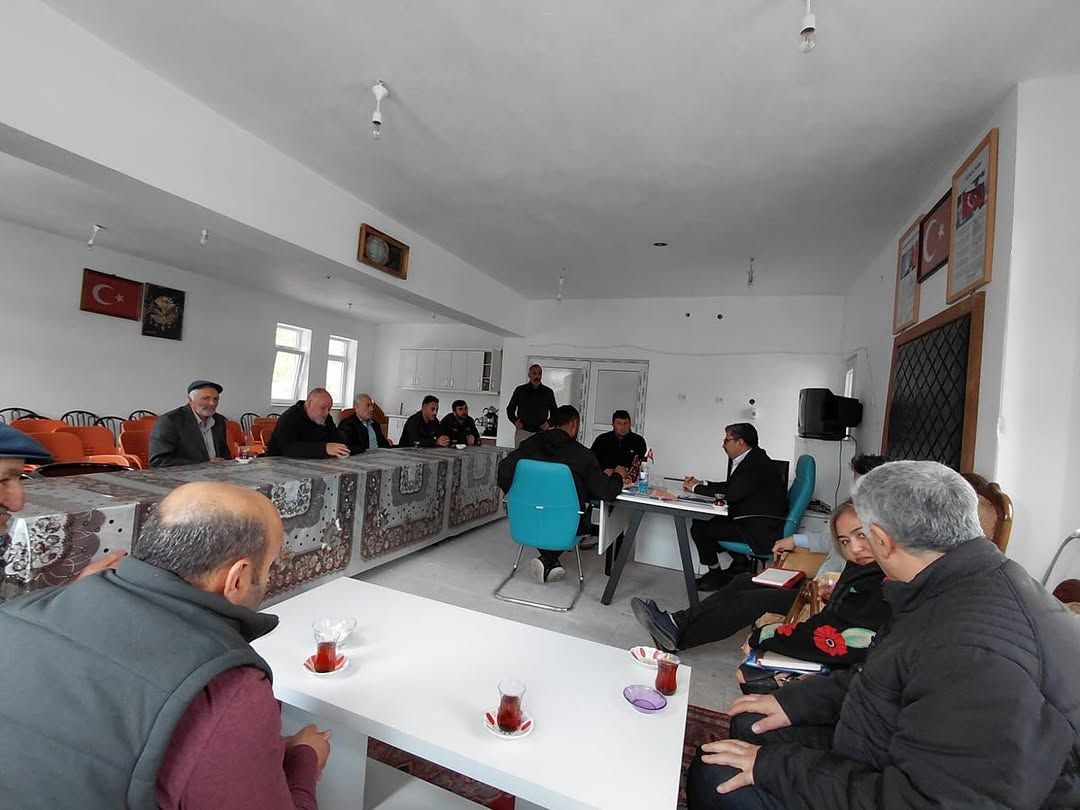 Toplantı salonunda masalar çevresinde oturan yetişkinler, çay bardakları ve duvarda Türk bayrağı görülüyor.
