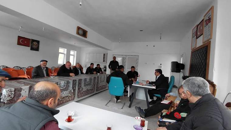 Toplantı salonunda masalar çevresinde oturan yetişkinler, çay bardakları ve duvarda Türk bayrağı görülüyor.