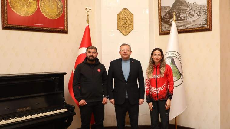 Bir odada üç kişi, Türk bayrağı ve başka bir bayrağın yanında, piyano önünde yan yana duruyor.