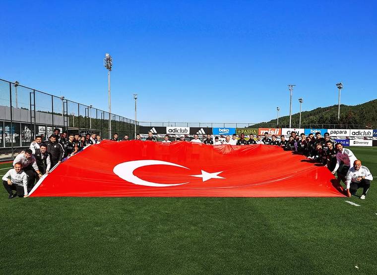 Futbol sahasında çok sayıda kişi, yere serilmiş büyük Türk bayrağının arkasında sıralanıyor.