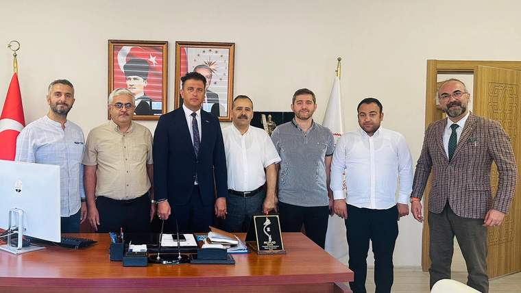 Ofis ortamında, Türk bayrağı ve duvar portreleri önünde duran yedi kişi; masada bilgisayar ve plaket bulunuyor.