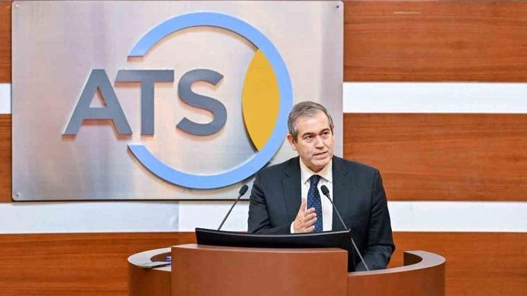 Kürsüde konuşan bir erkek, arkasındaki panoda büyük ATS harfleri ve dairesel bir logo önünde duruyor.