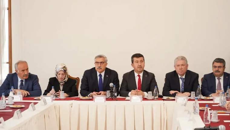 Bir toplantıda oturan altı kişi, masanın etrafında yer alıyor. Önlerinde su şişeleri ve tabaklar var.