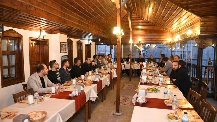 Ahşap tavanlı restoranda, uzun masalarda yemek yiyen ve oturan kalabalık bir grup görülüyor.