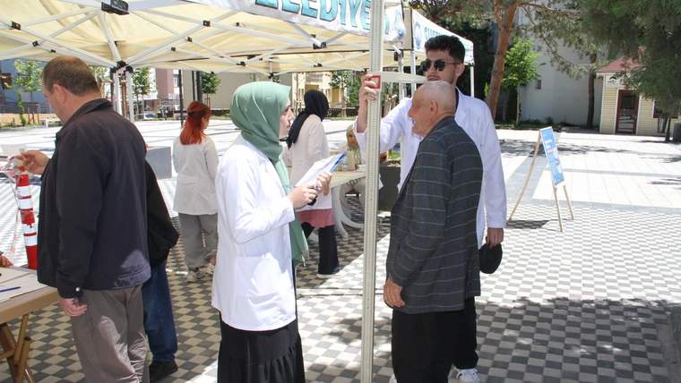 Açık alandaki stantta beyaz önlüklü kişiler, bir erkeğin tartı üzerinde durduğu sırada konuşuyor.