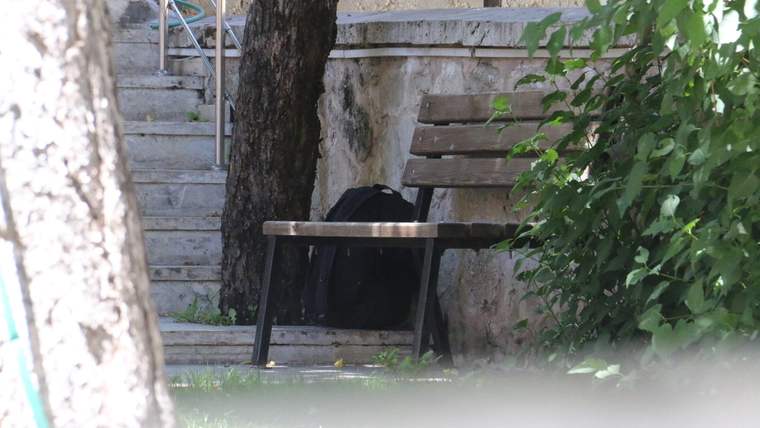 Ağaçların yanında, ahşap bankın altında siyah bir çanta ve arka planda taş basamaklar görülüyor.
