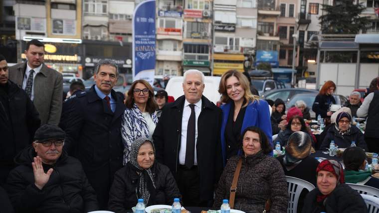 Açık alanda yemek masası çevresinde oturan ve ayakta duran kalabalık grup kameraya bakıyor.