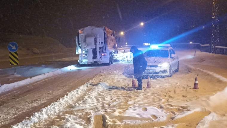 Gece karla kaplı yolda mavi ışıkları yanan bir araç, kamyon ve konilerin yanında duran kişi görülüyor.