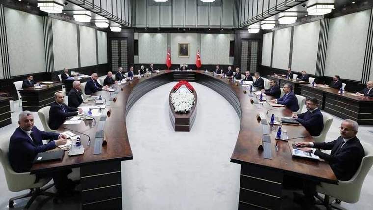 U biçimli toplantı masaları çevresinde çok sayıda kişi oturuyor; salonun ortasında çiçek aranjmanı bulunuyor.