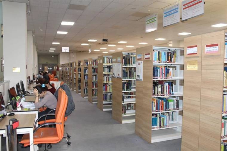 Kütüphane içinde kitap rafları ve bilgisayar başında çalışan kişiler görünmekte.