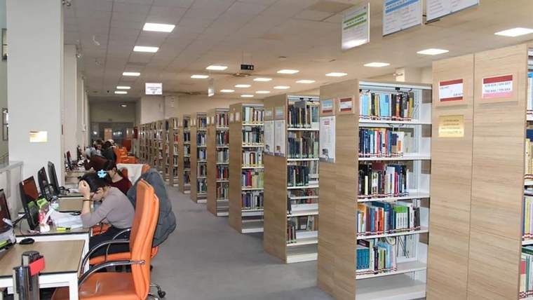 Kütüphane içinde kitap rafları ve bilgisayar başında çalışan kişiler görünmekte.