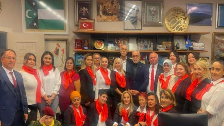 Kırmızı şal takan kadınlar, bir grup halinde poz veriyor; arka planda Türk bayrağı ve çeşitli tablolar var.