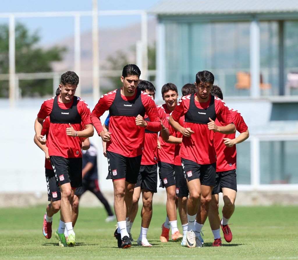 Kırmızı antrenman formaları ve siyah şortlar giyen futbolcular, çim sahada birlikte koşuyor.