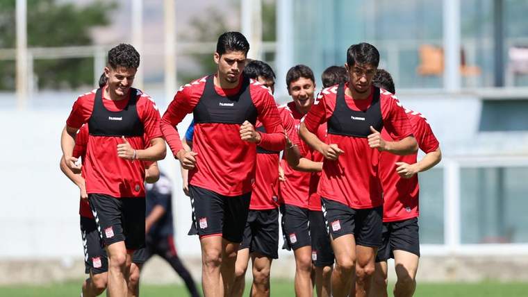 Kırmızı antrenman formaları ve siyah şortlar giyen futbolcular, çim sahada birlikte koşuyor.