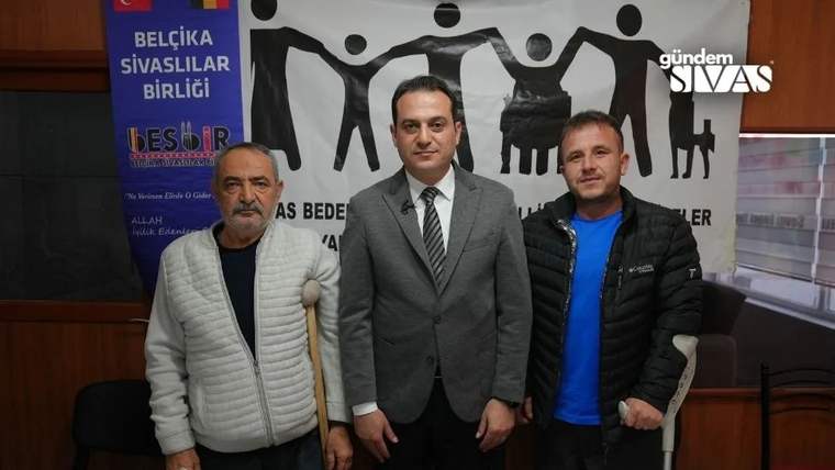 Toplantı salonunda üç erkek yan yana duruyor; ortadaki takım elbiseli, sağdaki koltuk değneği kullanıyor.