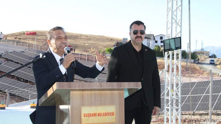 Güneş panelleri önünde kürsüde konuşan kişi ve yanında duran kişi, Suşehri Belediyesi tabelasının önünde.