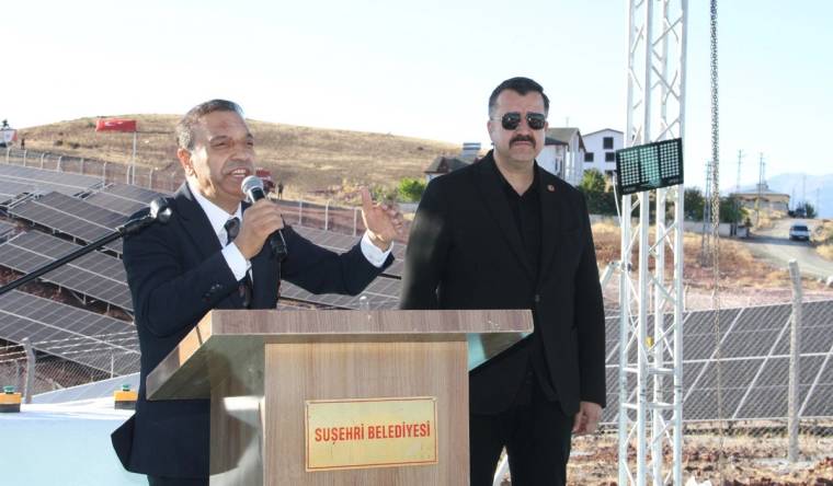 Güneş panelleri önünde kürsüde konuşan kişi ve yanında duran kişi, Suşehri Belediyesi tabelasının önünde.