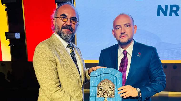 Bir kişi, elinde mavi bir plaket tutarken diğer kişiyle birlikte poz veriyor.