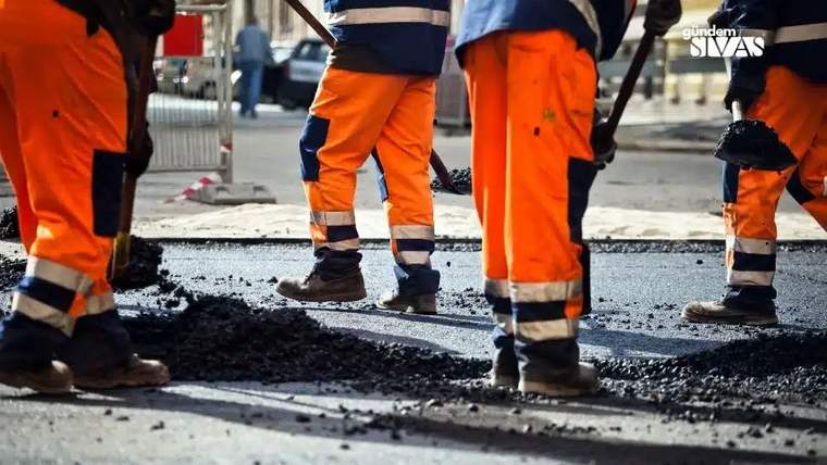 Asfalt döken işçilerin turuncu ve mavi iş kıyafetleriyle çalıştığı bir sokak.