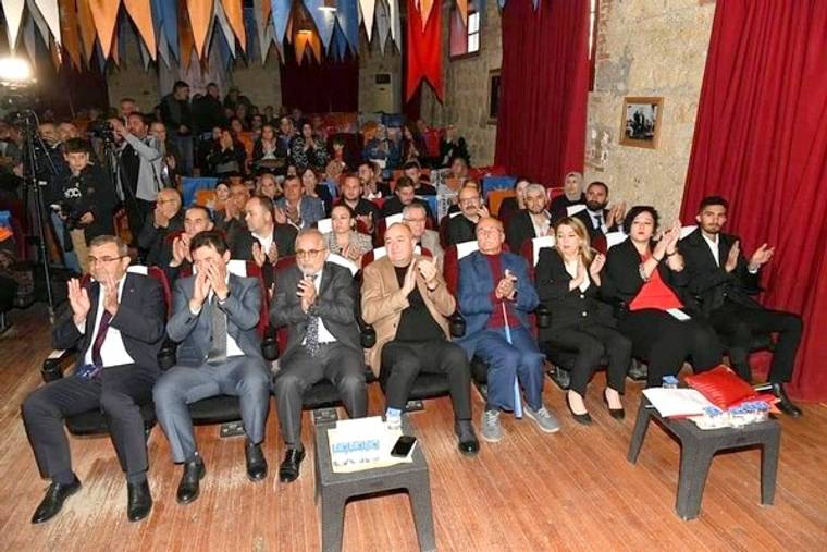 Taş duvarlı salonda sıralar halinde oturan kalabalık, ön sırada ellerini çırparken görülüyor.
