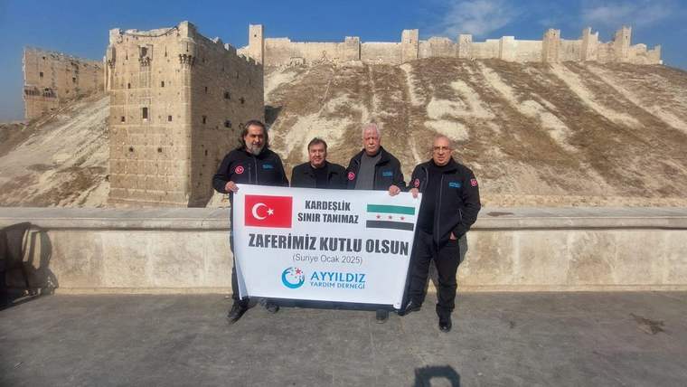 Dört kişi, arka planda tarihi taş yapı önünde üzerinde Türk bayrağı ve yazılar bulunan pankart tutuyor.
