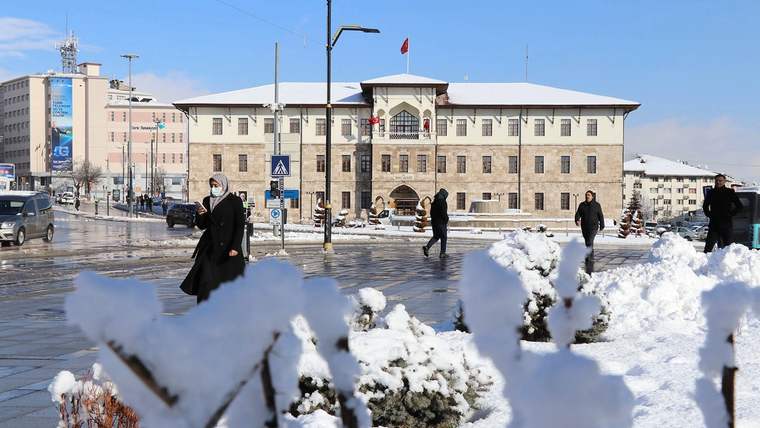 Karla kaplı bir alanda yürüyen insanlar, arka planda tarihi bir bina ve Türk bayrağı görünmekte.
