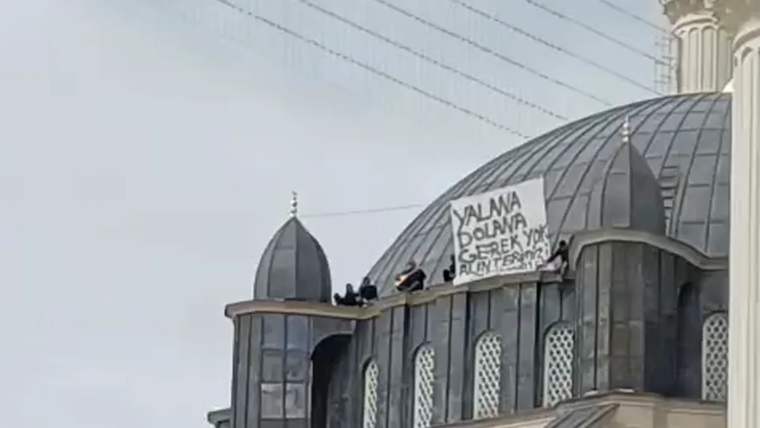 Kubbeli bir yapının çatısında birkaç kişi duruyor; korkulukta el yazılı bir pankart asılı.