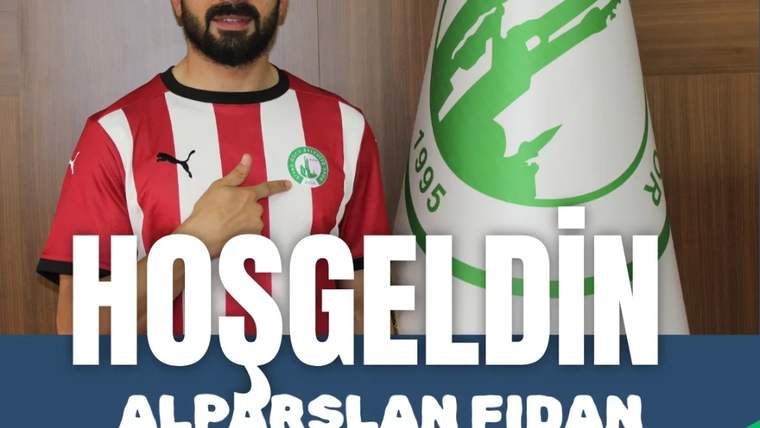 Kırmızı-beyaz çubuklu forma giyen sakallı bir kişi, yeşil-beyaz logolu bayrağın önünde poz veriyor.