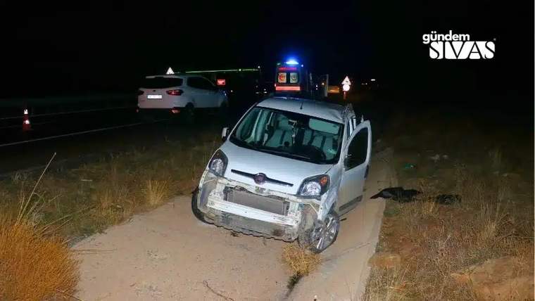 Gece, ön kısmı hasarlı beyaz bir minibüs yol kenarındaki toprak alanda duruyor; arkada mavi ışıklar görülüyor.